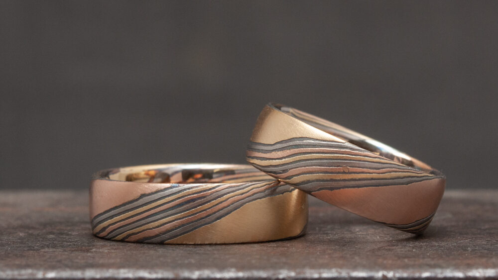 Zwei Mokume-Gane-Ringe mit geätzter Oberfläche aus Rotgold, Gelbgold, Palladium und geschwärztem Silber. Die Linien fließen diagonal, als wären sie vom Feuer gezeichnet.